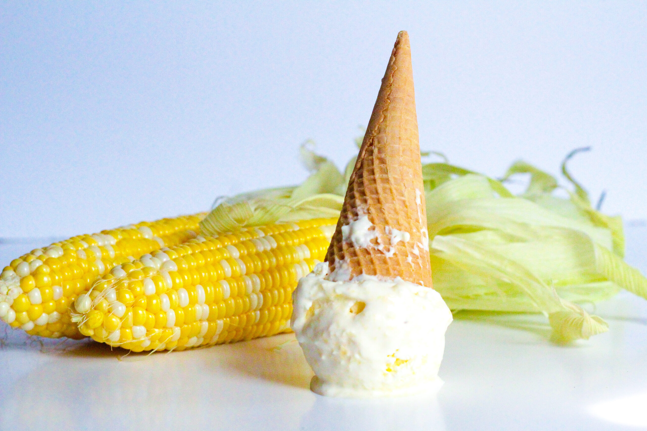 Side view of a scoop of sweet corn ice cream on a white surface with a cone going up from the scoop. Two cobs of corn are in the background.