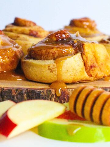 Side view of apples & honey cinnamon rolls on a round piece of wood with red and green apple slices in front of it and a honey stick resting on top