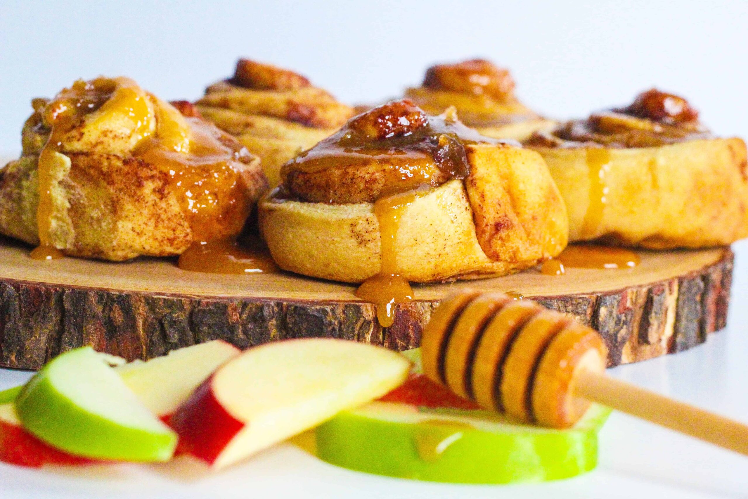 Side view of apples & honey cinnamon rolls on a round piece of wood with red and green apple slices in front of it and a honey stick resting on top
