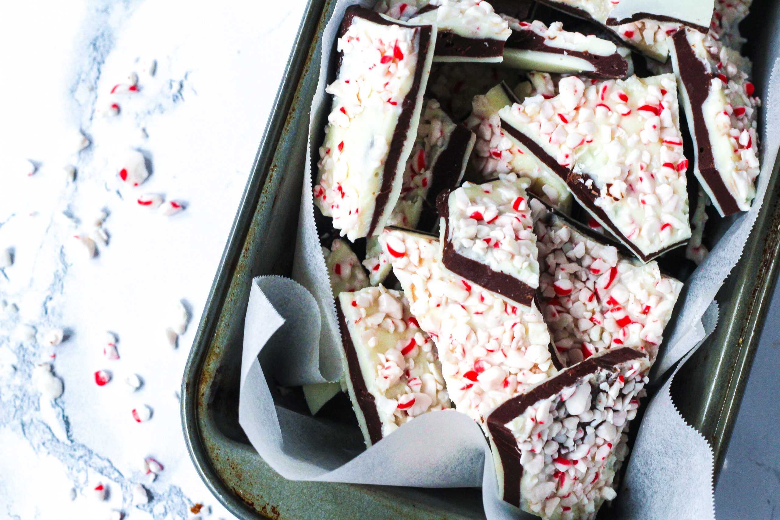 a silver loaf pan lined with parchment paper and filled with salted peppermint bark, coming in from the upper right of the frame at a slight angle. The white marbled surface is visible to the left with some crushed candy cane on it