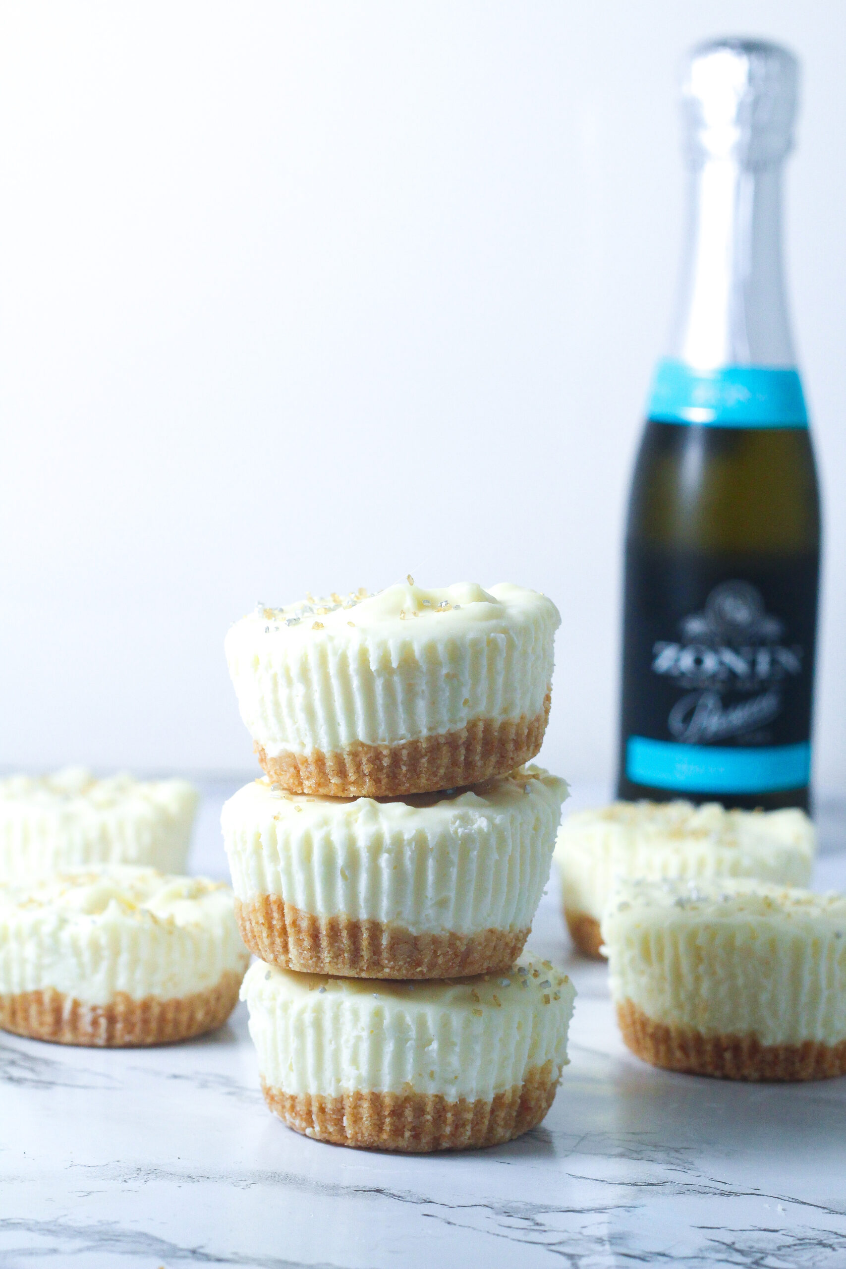 side view of a stack of 3 prosecco cheesecakes with other prosecco cheesecakes behind it and a mini bottle of prosecco in the back right of image