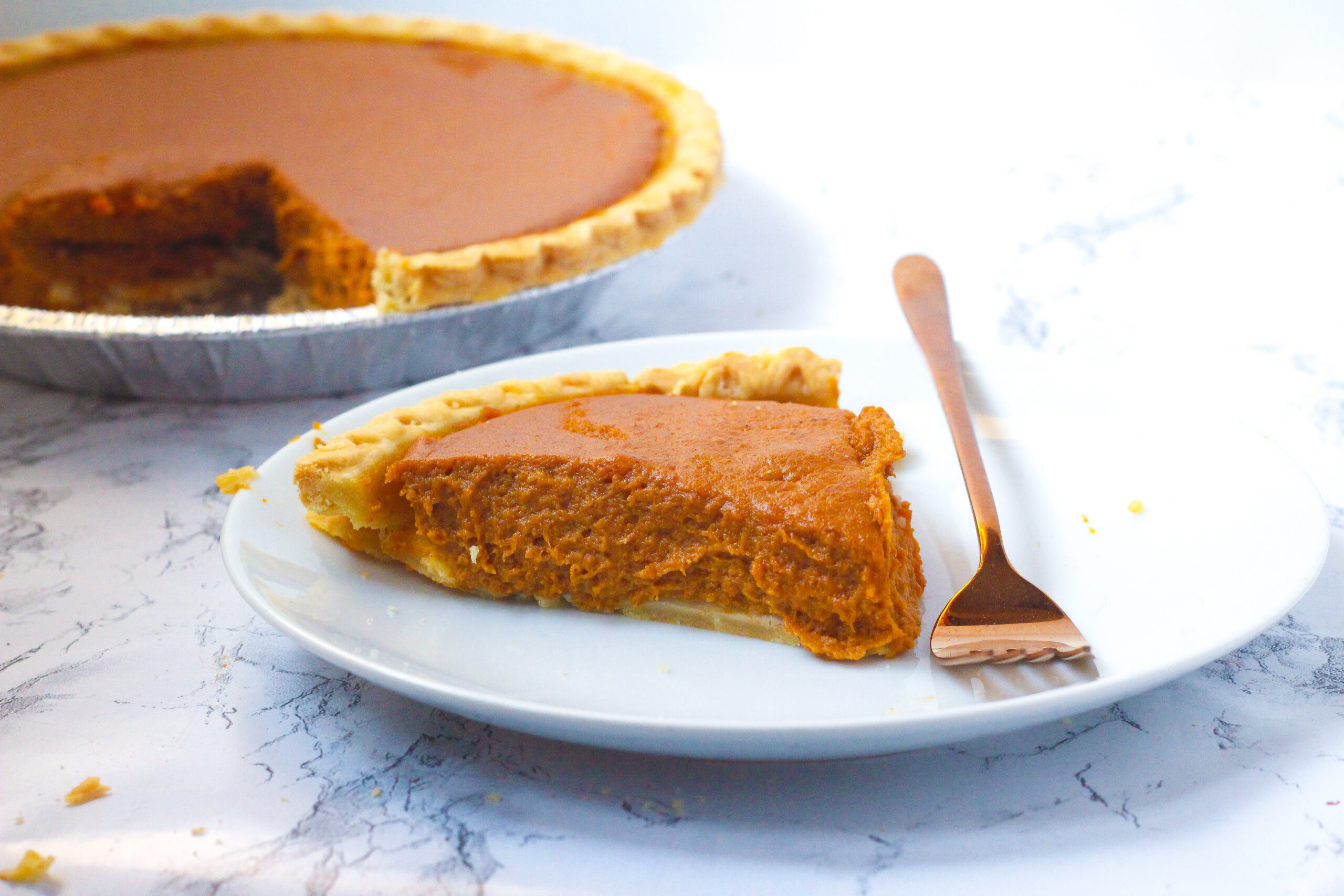 a slice of bourbon pumpkin pie on a round white plate with a rose gold fork on the plate to the right of the pie, with the fork prongs pointing toward the viewer. Behind the slice, off to the left is the remaining pie with the one slice missing.