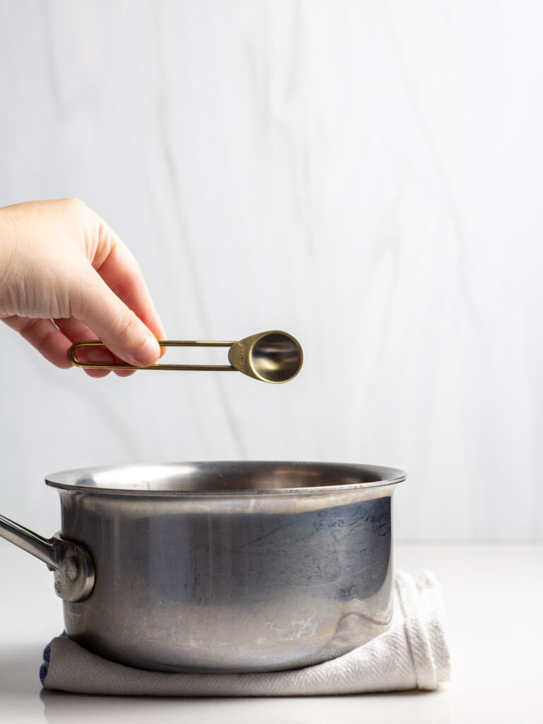 A hand holding a measuring spoon pouring peppermint extract into a saucepan.