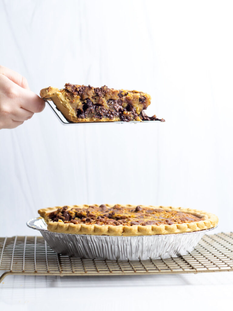 A hand holding a pie server with a slice of maple bourbon chocolate pecan pie over the rest of the pie.