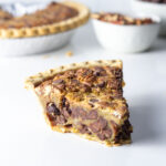 A slice of maple bourbon chocolate pecan pie, with the pie, pecans, and chocolate chips in the background.