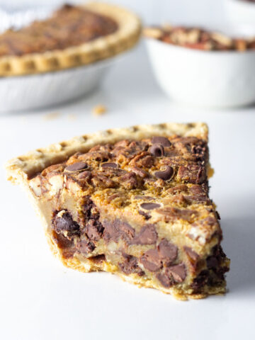 A slice of maple bourbon chocolate pecan pie, with the pie, pecans, and chocolate chips in the background.
