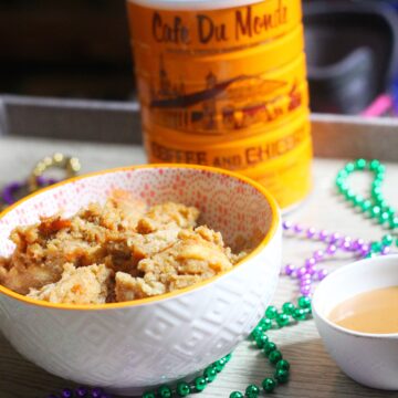 Cafe au Lait Bread Pudding in a white bowl with an orange rim next to a small white bowl of cafe au lait sauce, all surrounded by mardi gras beads, with a Cafe du Monde coffee can in the background