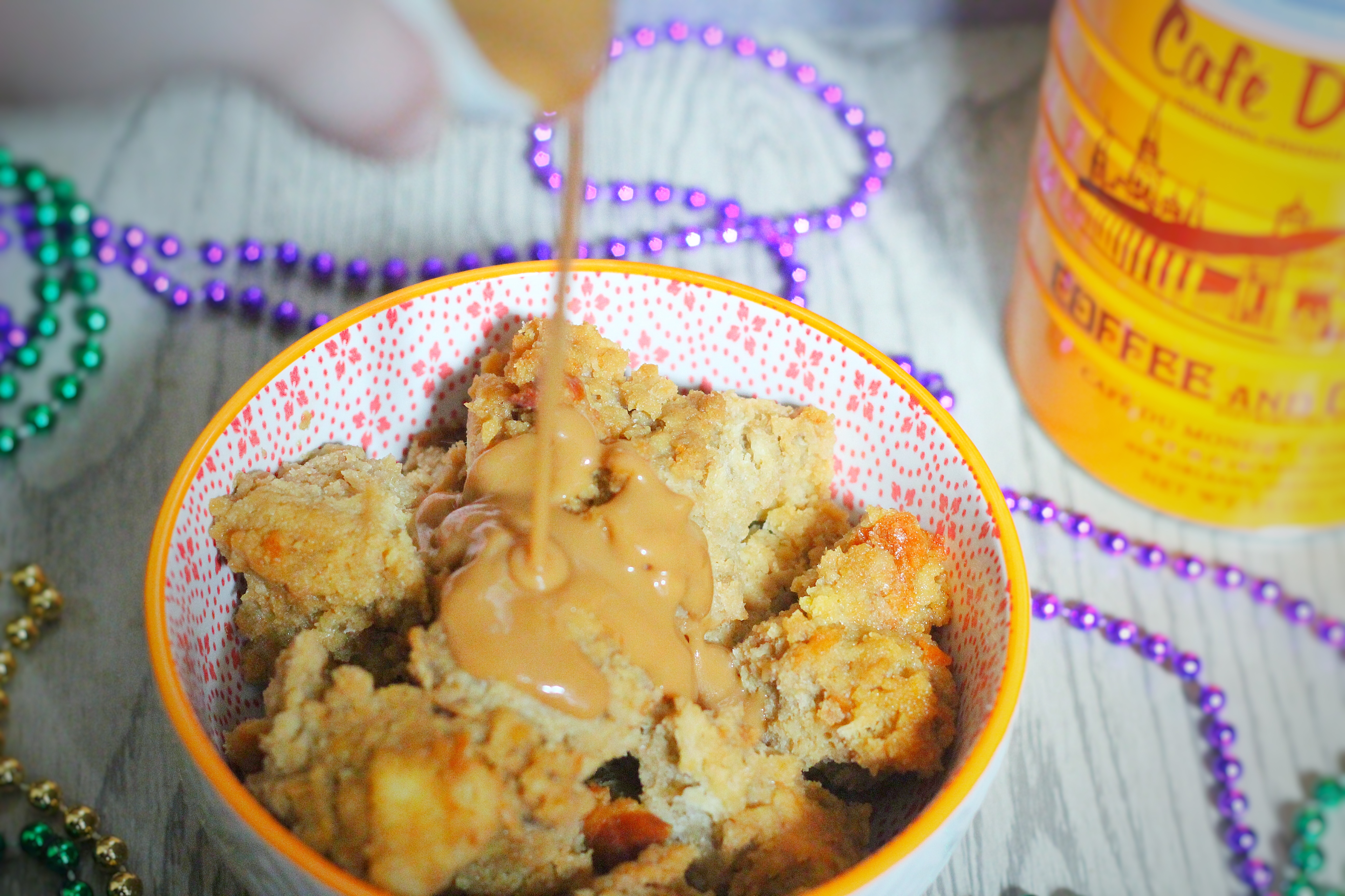 Cafe au Lait Bread Pudding in a white bowl with an orange rim surrounded by mardi gras beads, and a stream of cafe au lait glaze pouring onto it, with a Cafe du Monde coffee can in the background