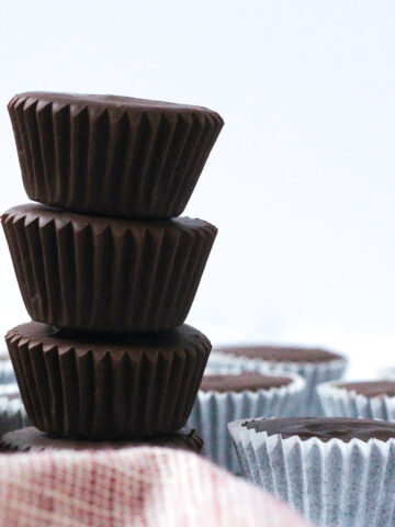 Side view of a stack of 4 dark chocolate peanut butter cups to the right of a red and white napkin. To the right of the stack are a single layer of peanut butter cups in white paper wrappers.
