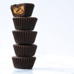 side view of a stack of 5 peanut butter cups with a bite taken out of the top one and the peanut butter filling exposed. The stack is surrounded by a white background.