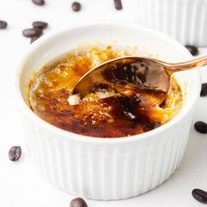 A rose gold spoon breaking through the caramelized sugar topping of a coffee creme brûlée in a white ramekin surrounded by coffee beans with more creme brûlées in the background.
