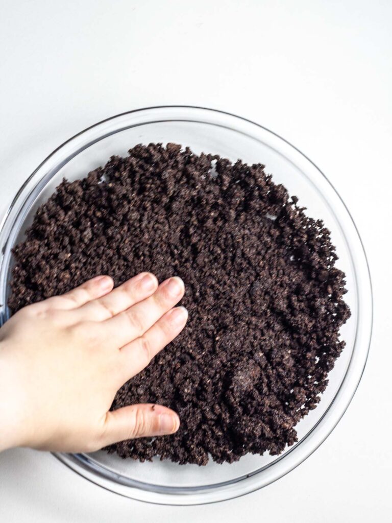 A hand pressing an Oreo crumb crust into a glass pie dish.
