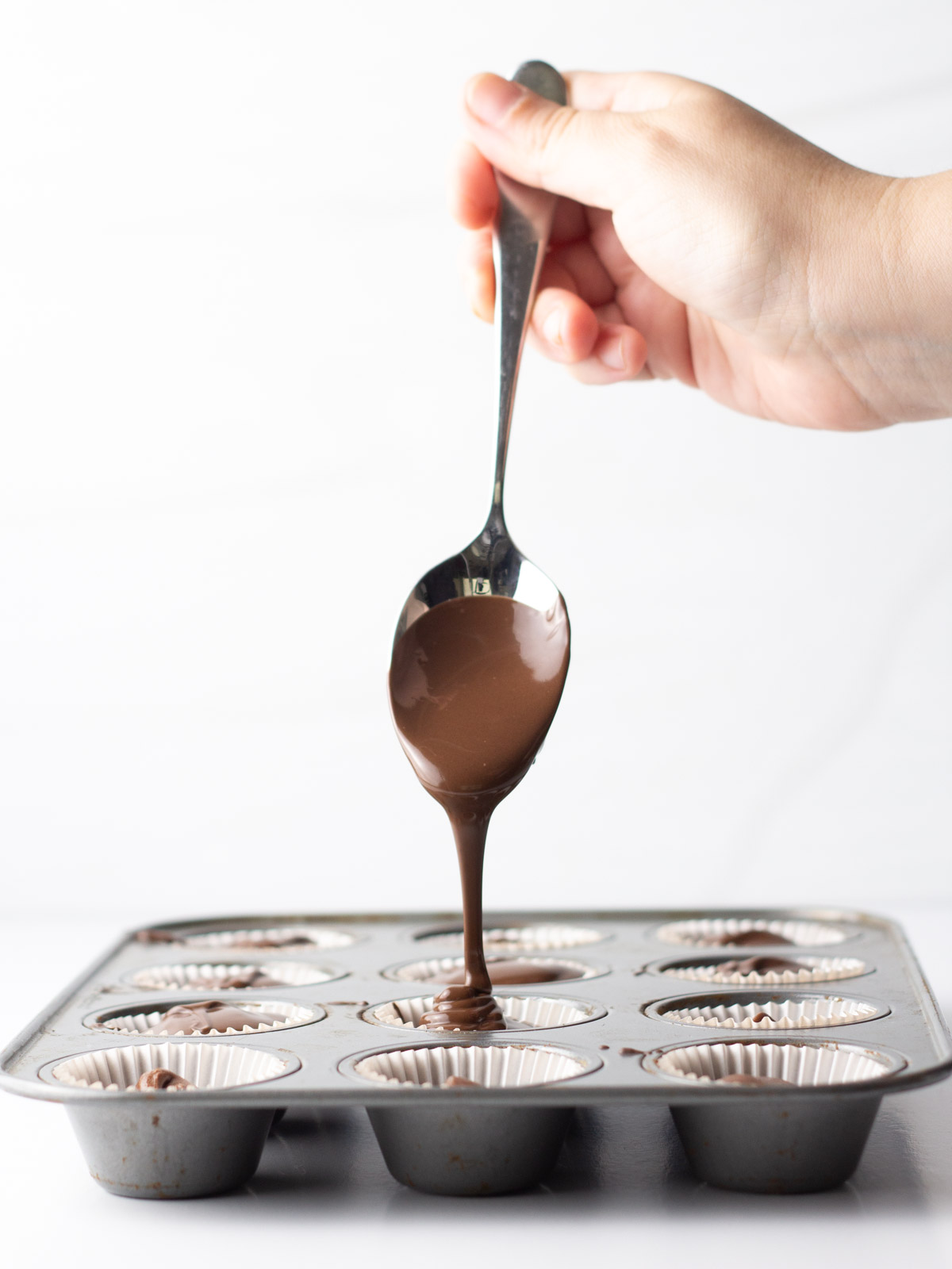 A hand holding a spoon with chocolate drizzling into the filled muffin tin.
