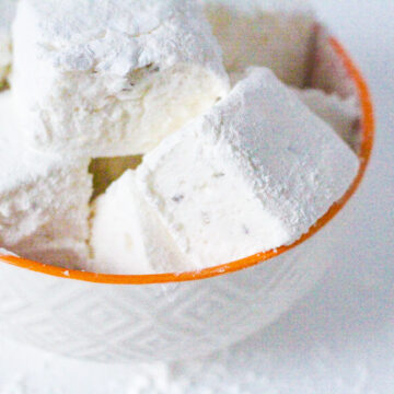 Angled top down view of a white textured bowl with an orange rim filled with lemon lavender marshmallows. The bowl is sitting slightly to the left of the frame and is cut off in the top left corner of the frame.