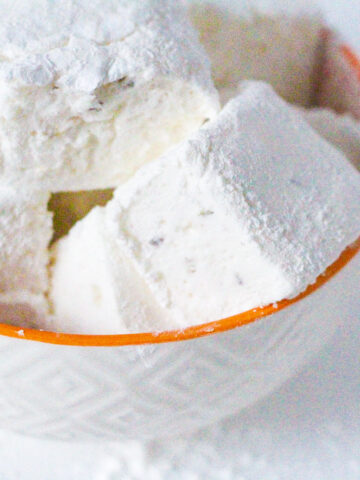 Angled top down view of a white textured bowl with an orange rim filled with lemon lavender marshmallows. The bowl is sitting slightly to the left of the frame and is cut off in the top left corner of the frame.