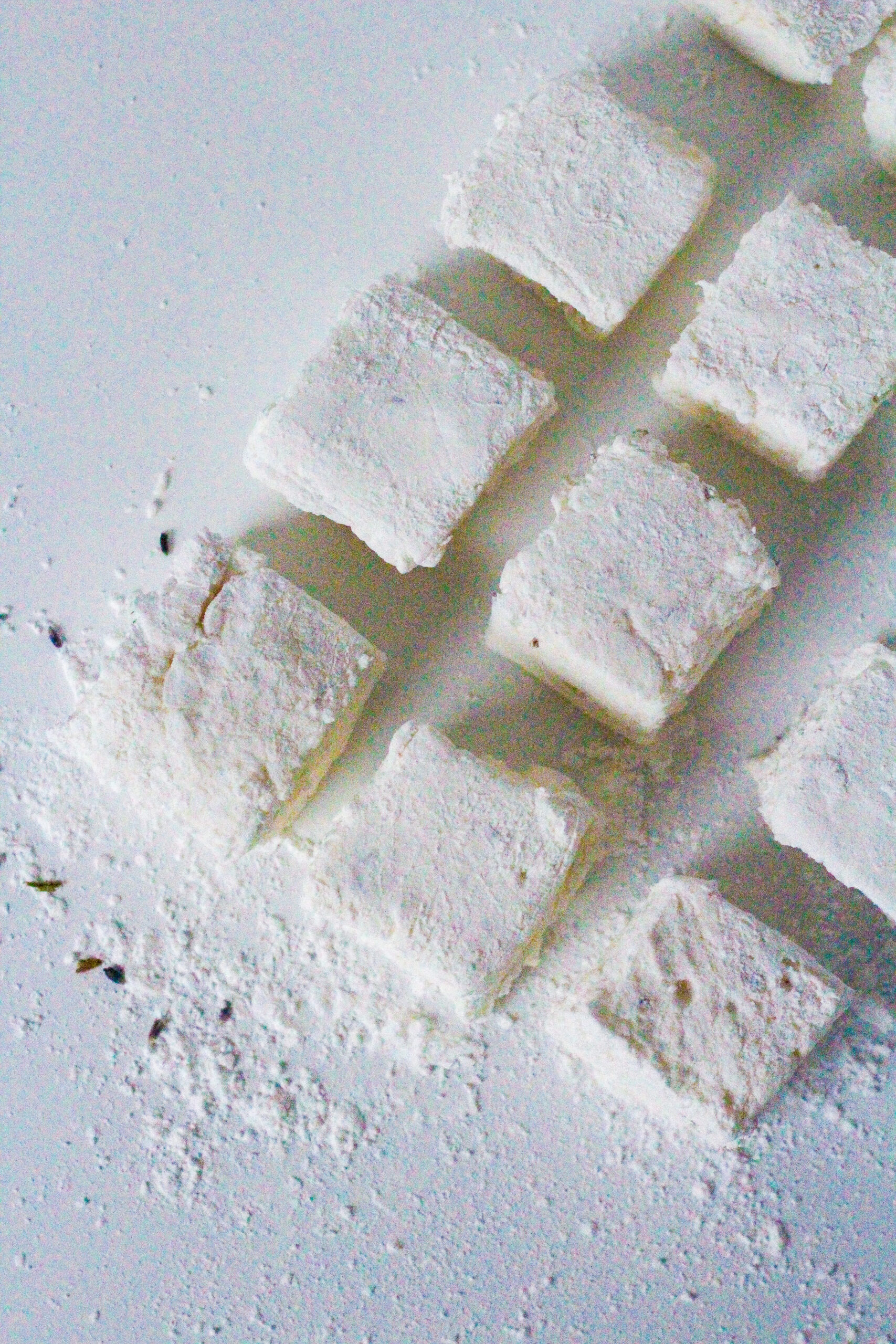 top down view of lemon lavender marshmallows arranged in a rectangular pattern with a bit of space between each marshmallow. The rectangle of marshmallows is coming into the frame from the top right corner at an angle so that going from the midleft side of the frame up to the top right side of the frame you can see three and a piece of a fourth marshmallows forming the left side of the rectangle. From that same center left marshmallow going down to the bottom right of the frame there are two more marshmallows forming the shorter bottom side of the rectangle. The center left marshmallow is surrounded by powdered sugar, corn starch, and lavender.