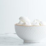 White textured bowl with orange rim filled with white meringues in front of a white background on a marbled surface