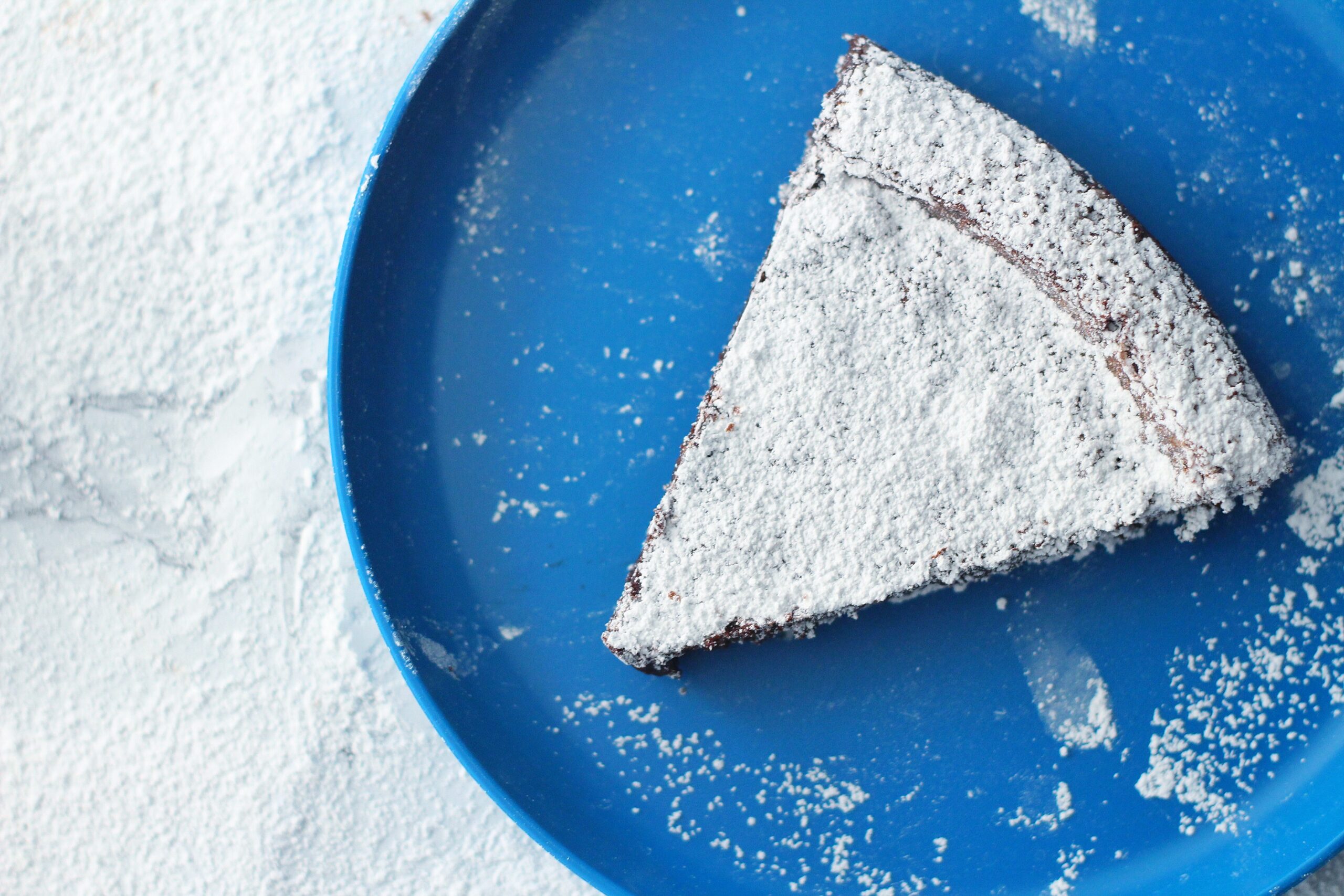 Slice of flourless chocolate cake on a round blue plate on top of a marbled surface covered in powdered sugar