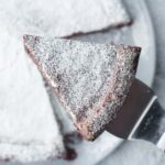 Once slice of flourless chocolate cake on a cake server held above the rest of the flourless chocolate cake blurred in the background. Both the slice and full cake are covered in powdered sugar. The full cake is sitting on a round, grey plate on top of a marbled surface