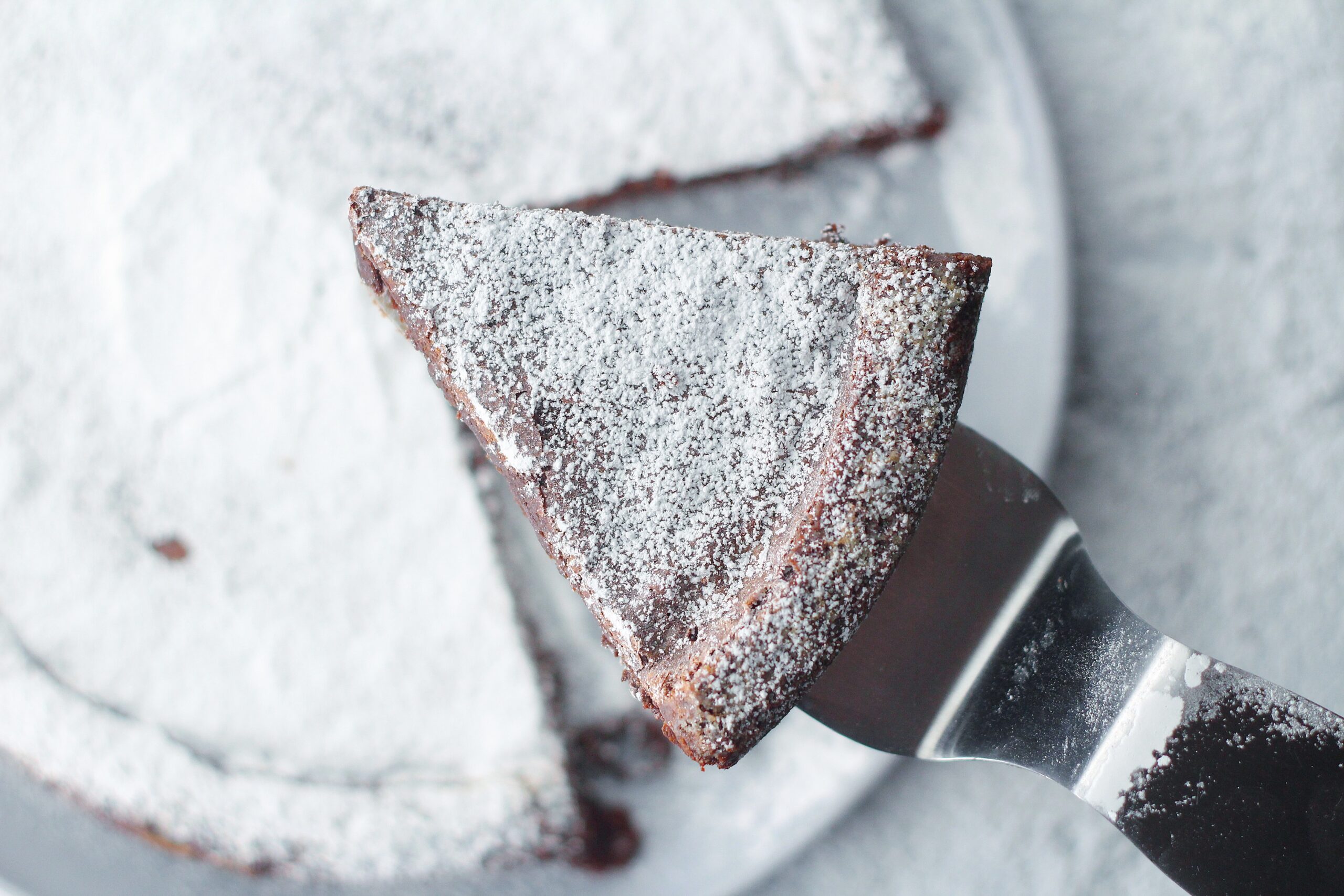 Once slice of flourless chocolate cake on a cake server held above the rest of the flourless chocolate cake blurred in the background. Both the slice and full cake are covered in powdered sugar. The full cake is sitting on a round, grey plate on top of a marbled surface