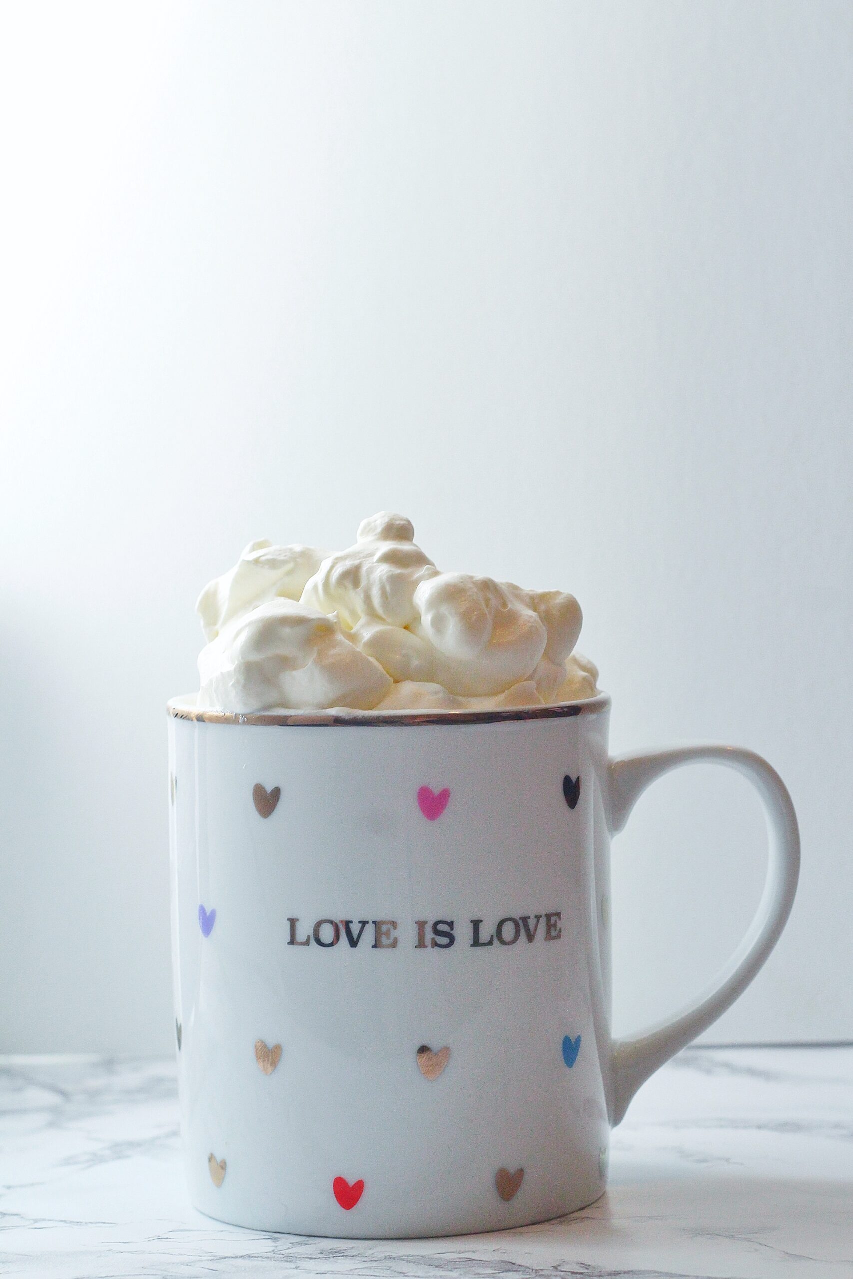 A white mug with a gold rim covered in multi-colored hearts that says love is love on the front with Bailey's whipped cream sitting on top