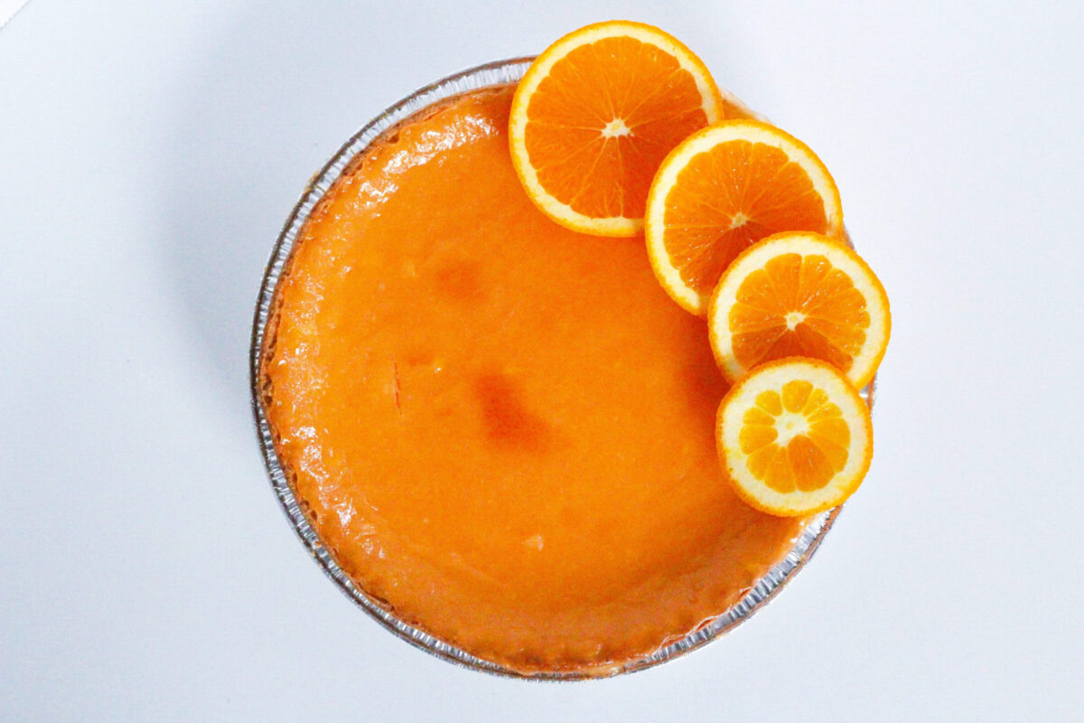 Top down view of a bright orange aperol spritz pie with four orange slices fanned along the right-side edge of the pie. The pie is sitting on a white surface.