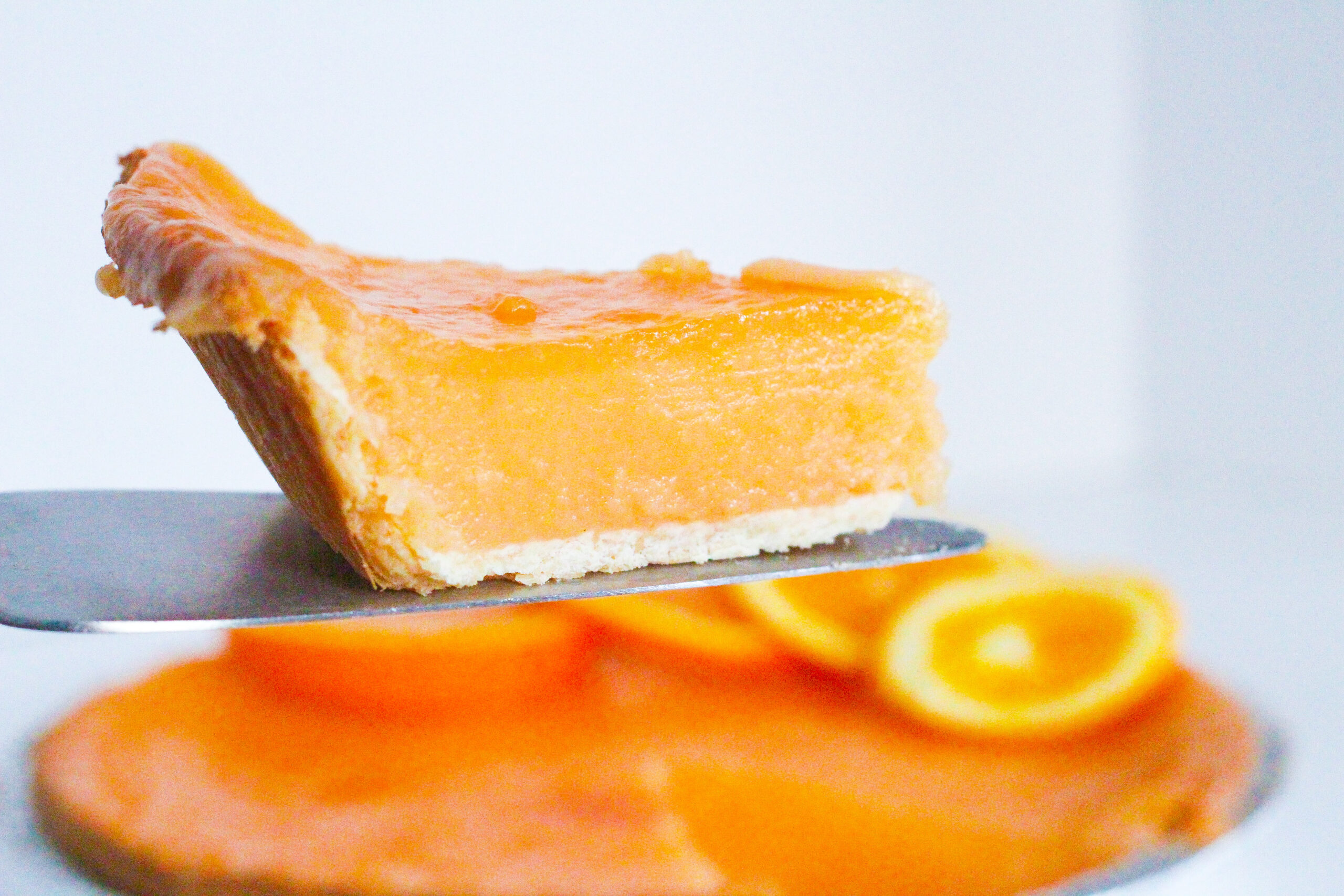 Side view of a slice of aperol spritz pie being held on a pie server above the rest of the aperol spritz pie. All on a white surface in front of a white background.