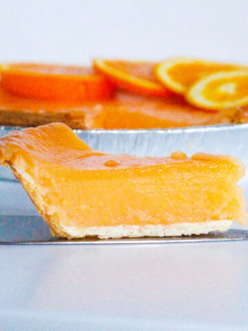 Side view of a slice of aperol spritz pie on a pie server sitting in front of the rest of the aperol spritz pie. All on a white surface in front of a white background.