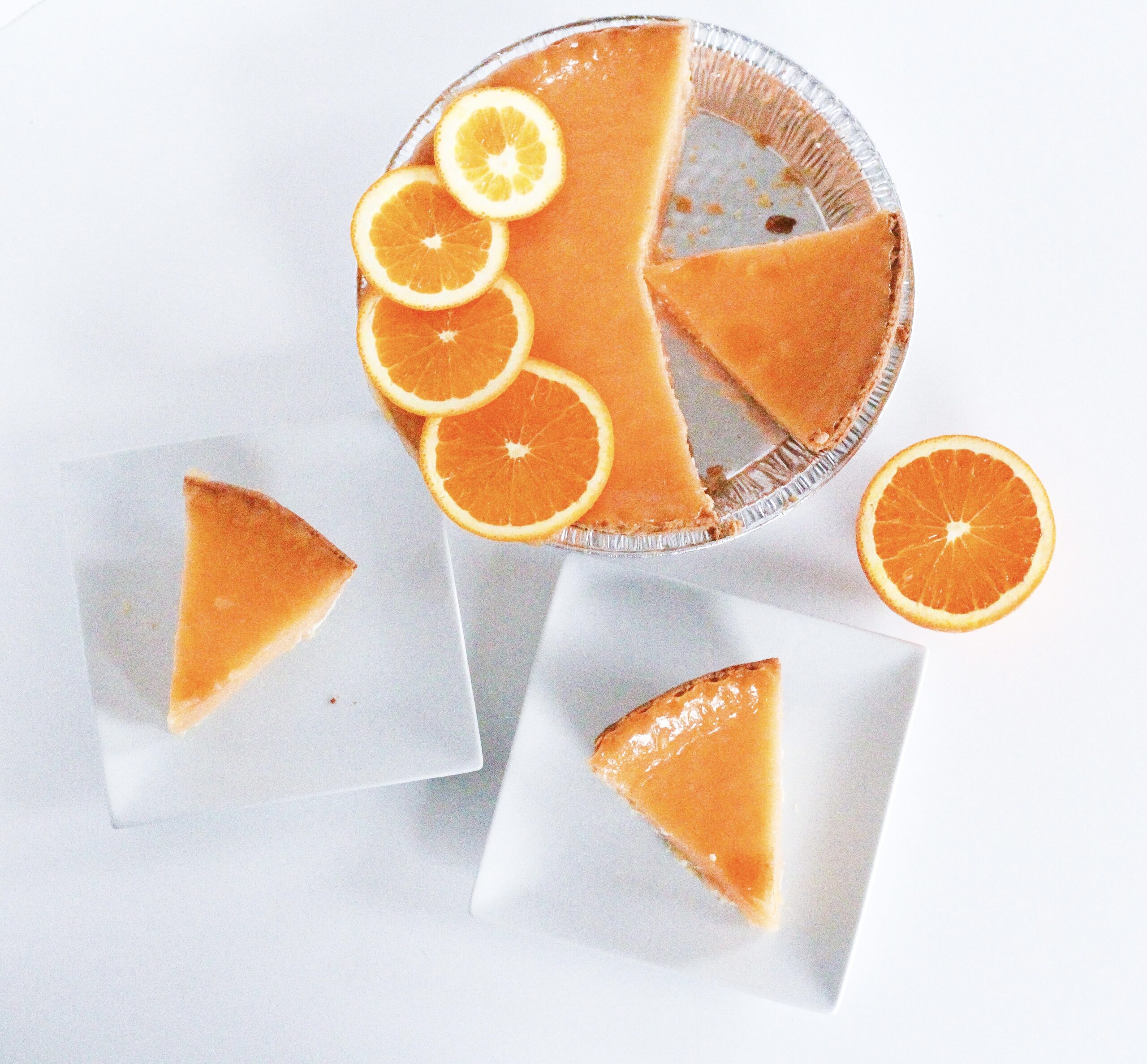 Top down view of the pie with three slices cut. One slice is in the open space in the pan where other slices have been removed. The other two slices are each on a small square white plate. There is also half an orange sitting next to the pie.