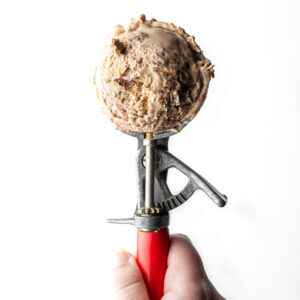 A hand holding the red handle of an ice cream scooper with a scoop of Nutella ice cream in the scooper.