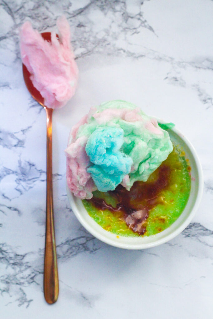 rose gold spoon with a spoonful of pink cotton candy vertically along the left hand side of the frame.  On the right just below center of the frame is a cotton candy creme brulee with blue and pink cotton candy on top. all on a white marbled surface.