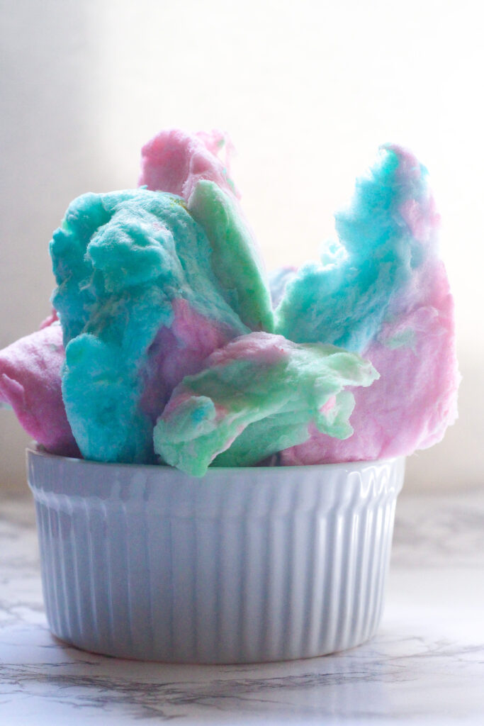 side view of a white ribbed ramekin with pink, blue, and green cotton candy piled on top like a sculpture! The ramekin in on a marbled surface and has a white background
