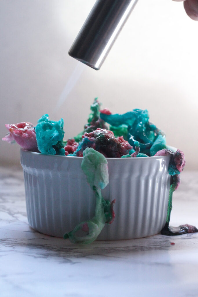 Side view of a white, ribbed ramekin with melting pink, blue, and green cotton candy on top, with a torch above it and a flame melting the cotton candy topping.