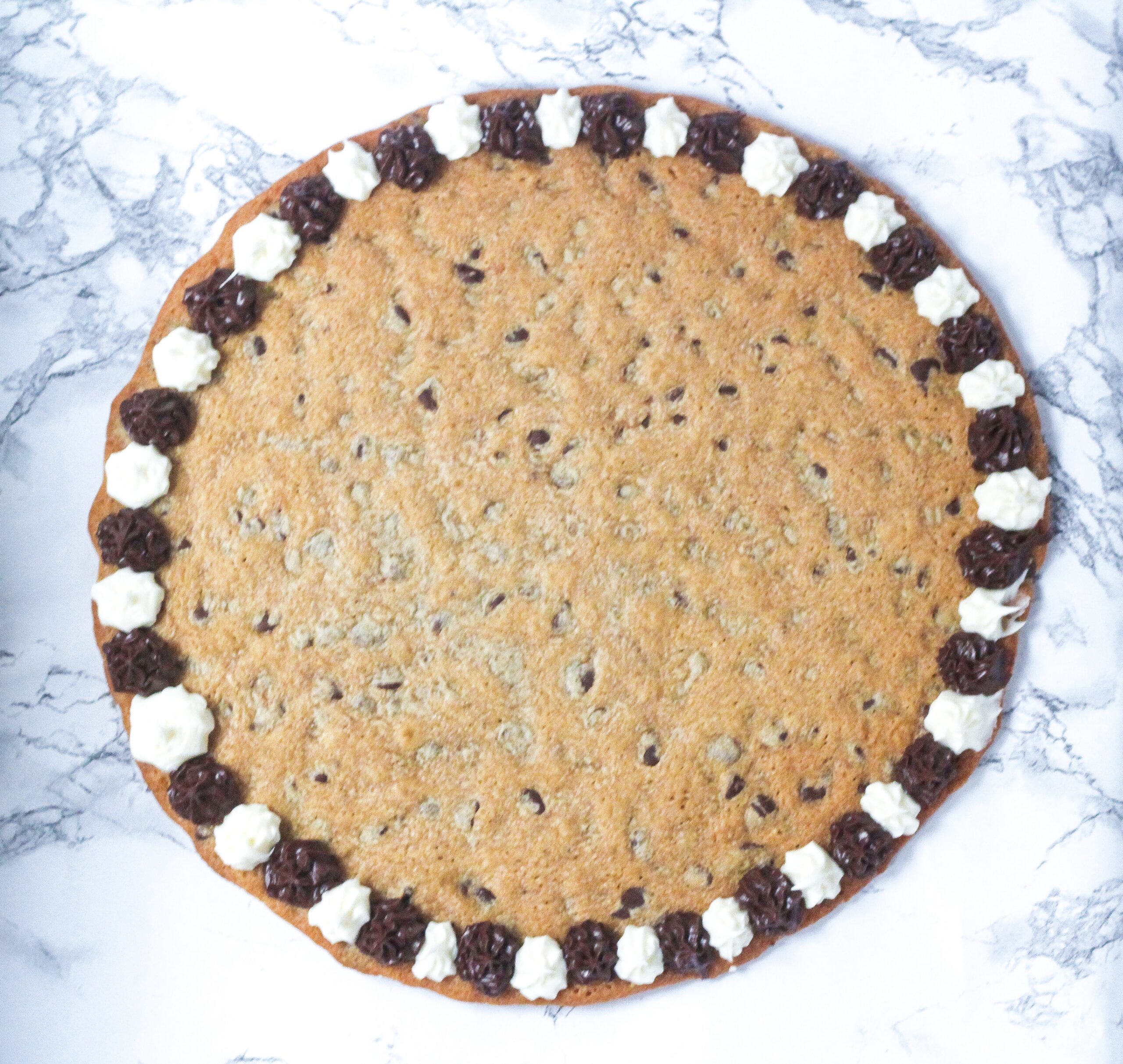 top down view of whole chocolate chip cookie cake with alternating brown and white frosting dots around the edge, on top of a marbled surface