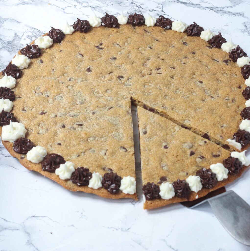 angled shot of a chocolate chip cookie cake with alternating brown and white frosting flowers around the outside of the cookie, with a slide in the bottom right of the cookie cut and sitting on a cake server, pulled slightly out, on a marbled surface