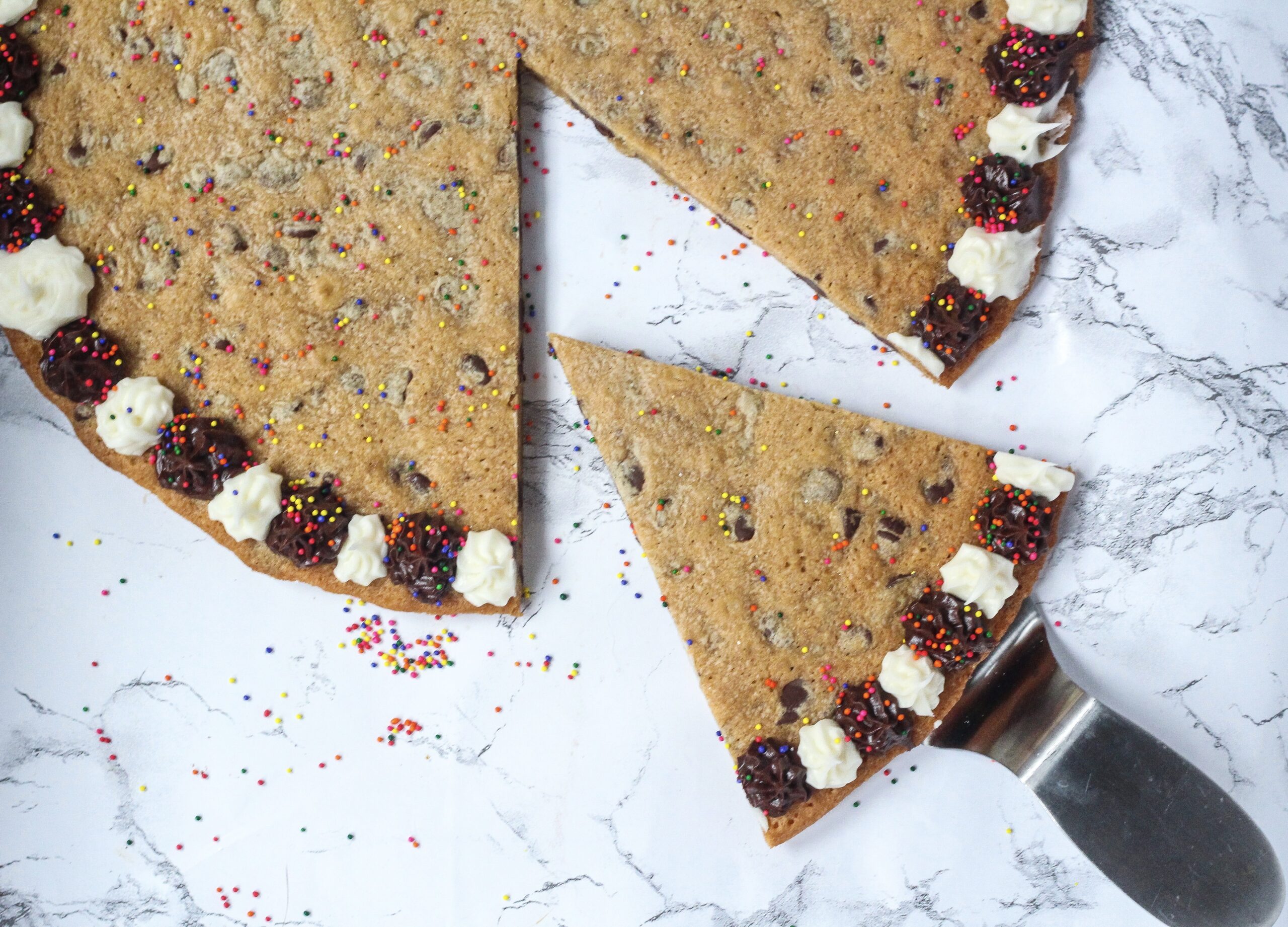 top down view of cookie cake with alternating brown and white frosting dots around the outside, and one slice pulled out part-way and angled. Multi-colored sprinkles are on the cookie as well as the marbled surface the cookie is on.