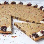 angled view of cookie cake with alternating brown and white frosting dots around the outside, and one slice pulled out part-way and angled. Multi-colored sprinkles are on the cookie as well as the marbled surface the cookie is on.