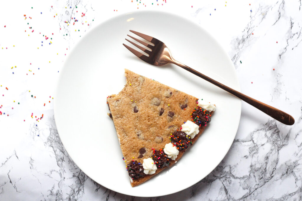 round white plate with a slice of chocolate chip cookie cake with the tip of the slice bitten off, and the alternating white and brown frosting dots along the bottom of the slice. To the right of the slice is a rose gold fork. The plate is on top of a white marbled surface.