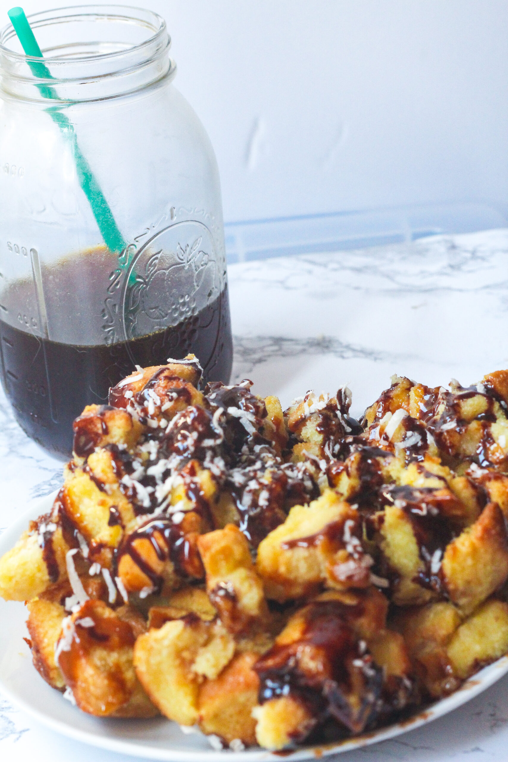 Caramel Chocolate Coconut Bread Pudding in a white baking dish on a white marbled surface with a mason jar of cold brew coffee in the back left corner with a green straw sticking out