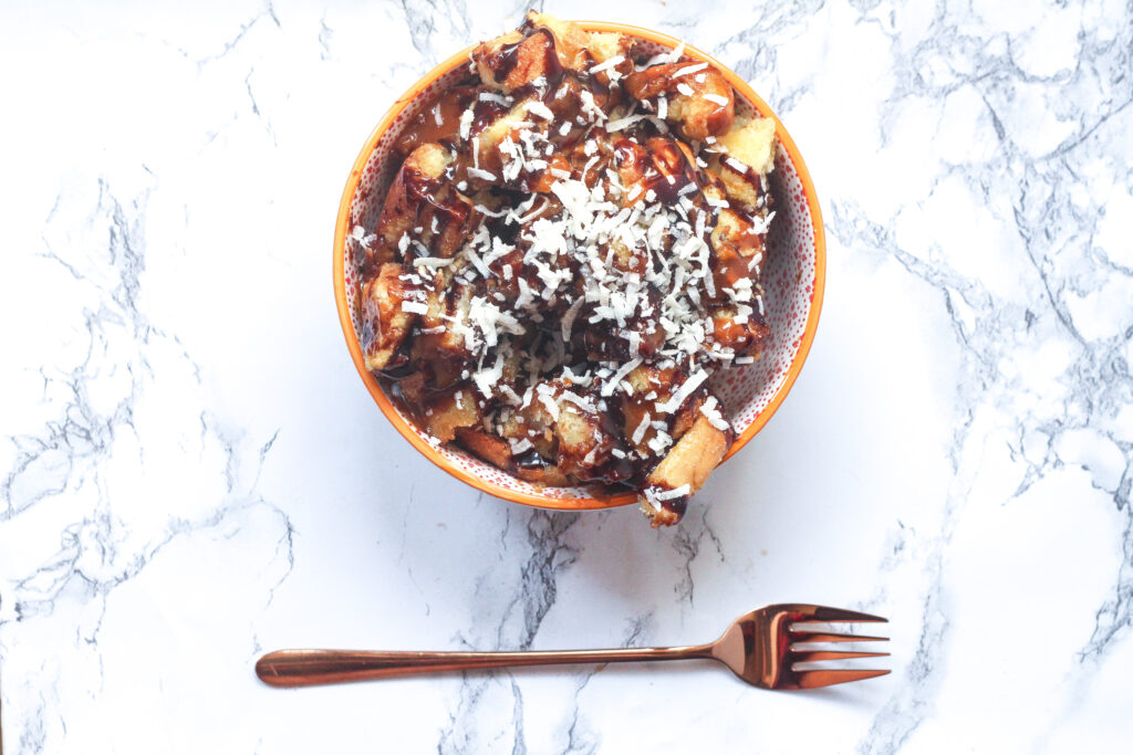 a bowl with an orange rim filled with caramel chocolate coconut bread pudding topped with chocolate sauce, caramel sauce, and shredded coconut in the center of the photo on top of a marbled surface, with a rose gold fork laying horizontally below the bowl