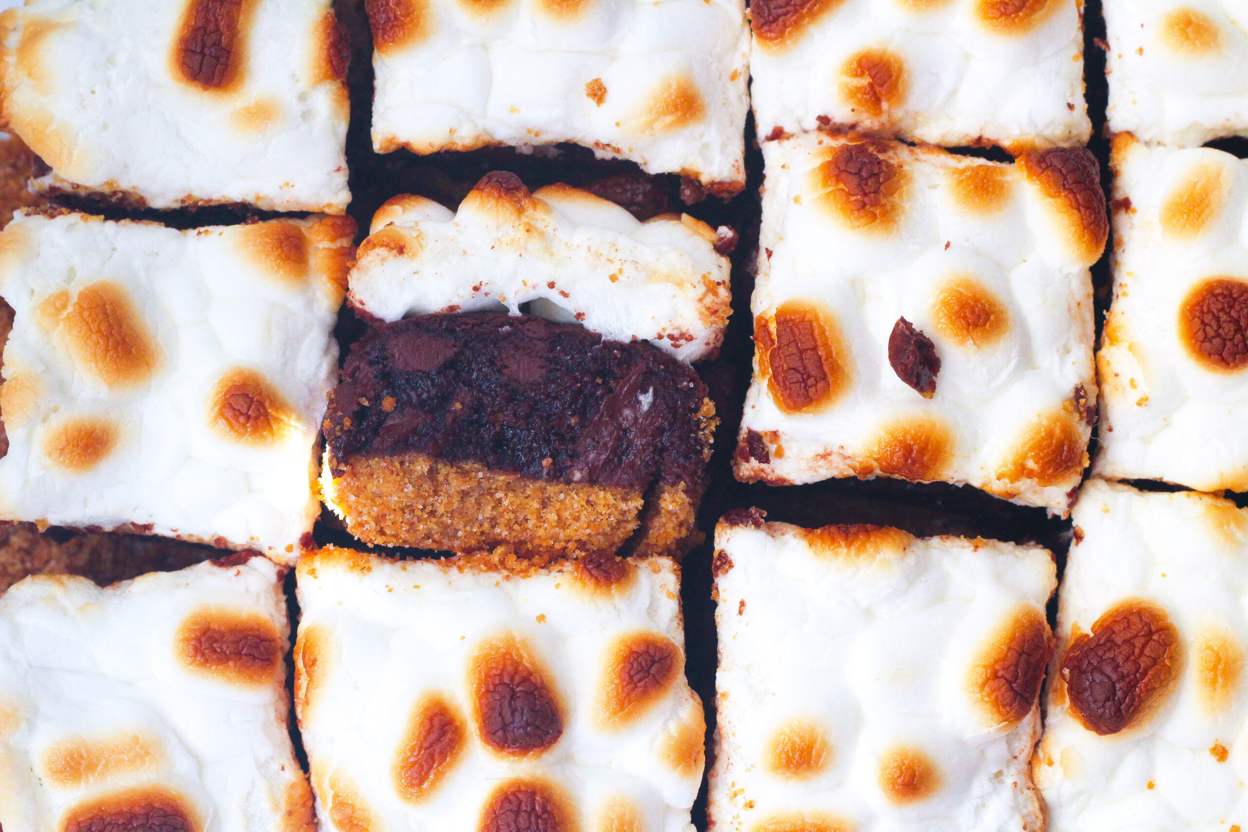 Top down view of the toasted marshmallow top of Bailey's S'mores Brownies, with one brownie turned sideways to see the three layers - the light brown graham cracker crust, the dark brown brownie in the center, and the white marshmallow topping