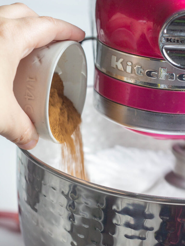 A hand pouring pumpkin pie spice into the running pink kitchenaid mixer.
