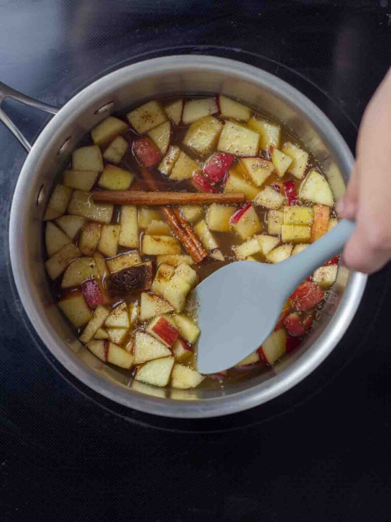 Chopped apples, cinnamon sticks, and spices in a pot on the stove, being stirred.