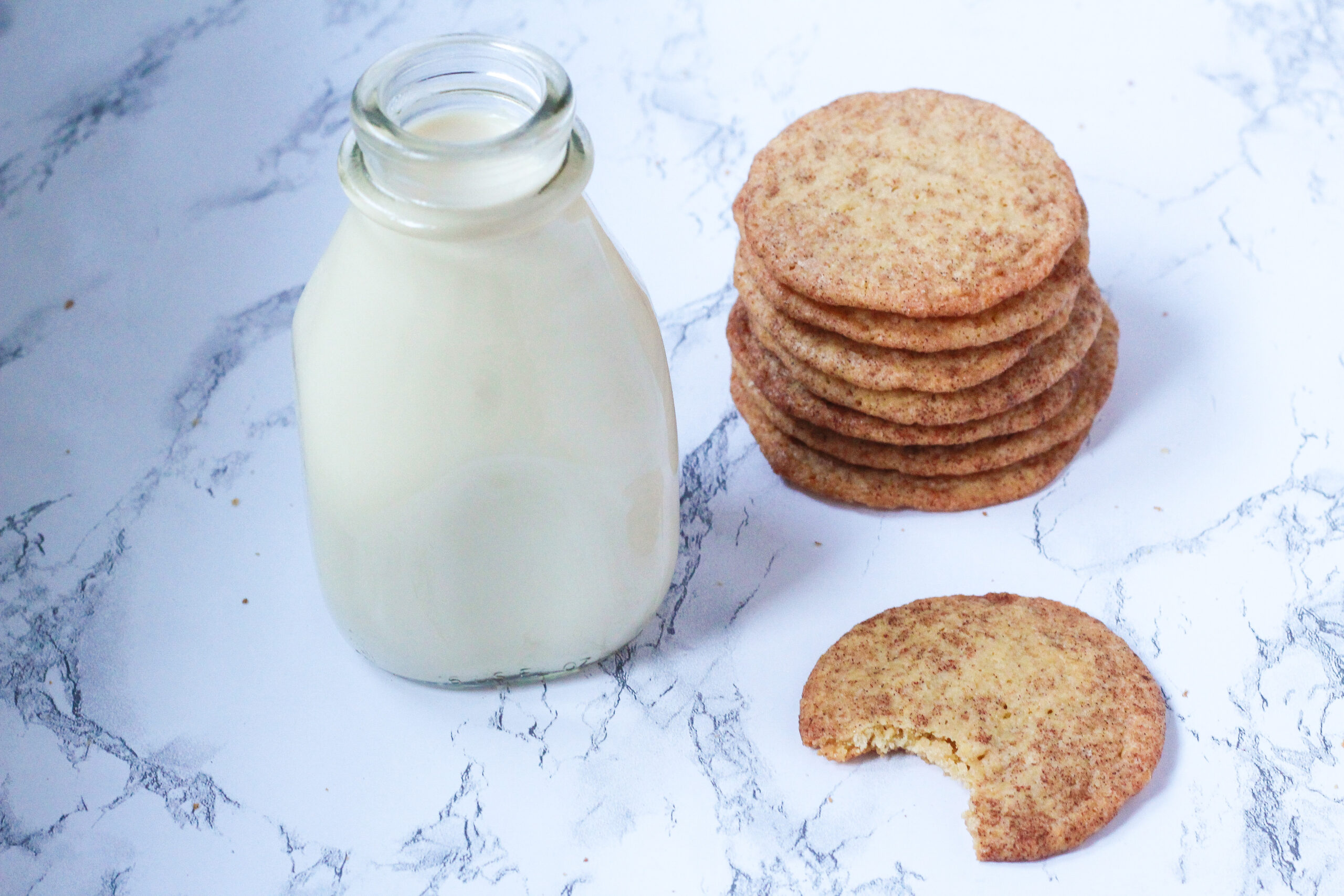 Stack of french toast cookies to the right of a glass milk jar, with a cookie with a bite out of it in front of the cookie stack, all on a marbled surface
