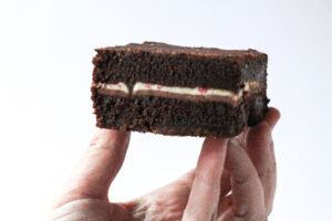 A hand in the bottom of the frame, cupped with the fingers pointing upwards holding a brownie on top of the fingers. The Peppermint mocha brownies are dark brown brownies with a thin strip of white in the center from the peppermint bark square