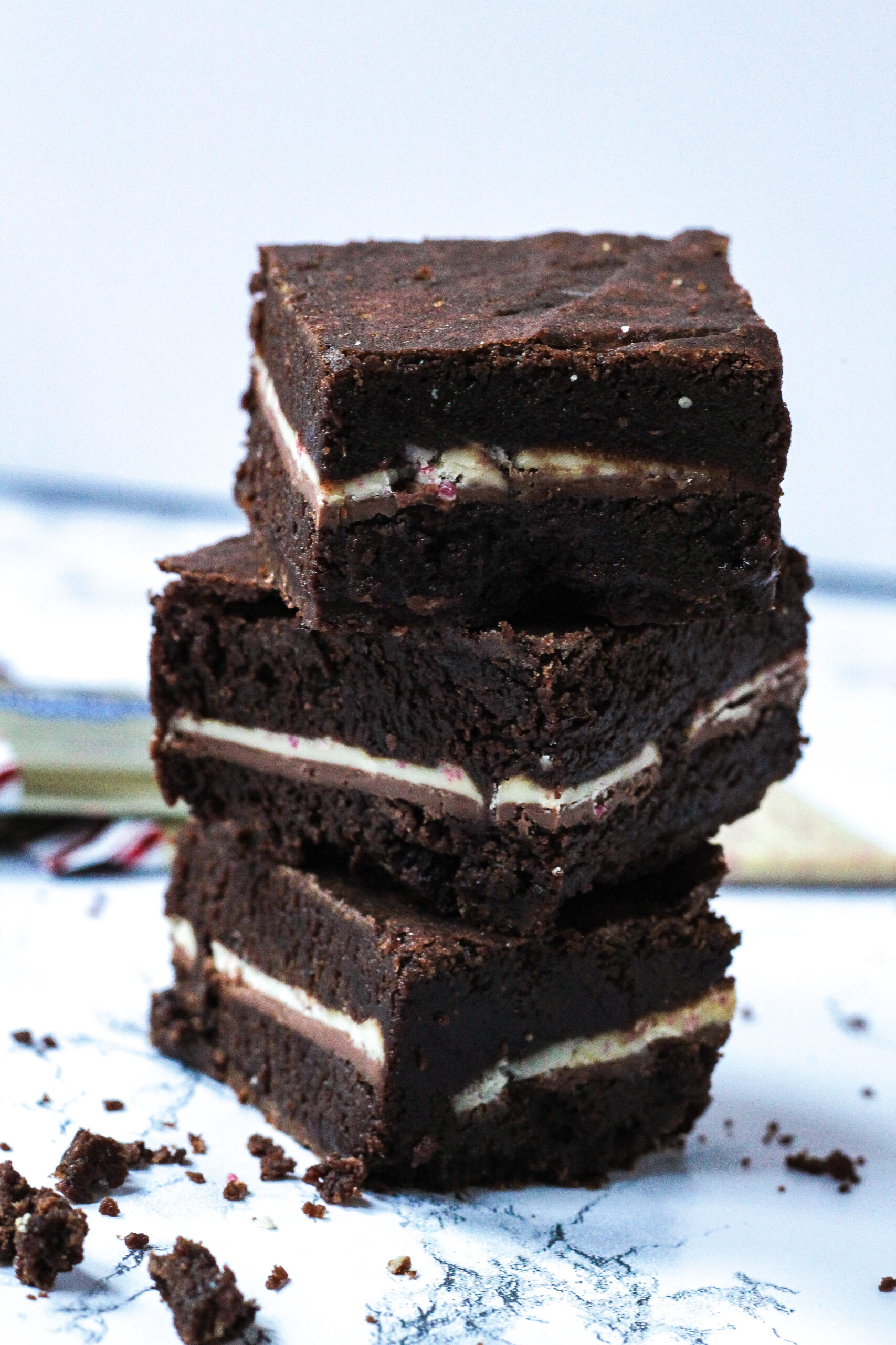 side view of a stack of three peppermint mocha brownies with crumbs on the white marbled surface around the stack. Barely visible behind the stack are some wrapped and unwrapped peppermint bark squares