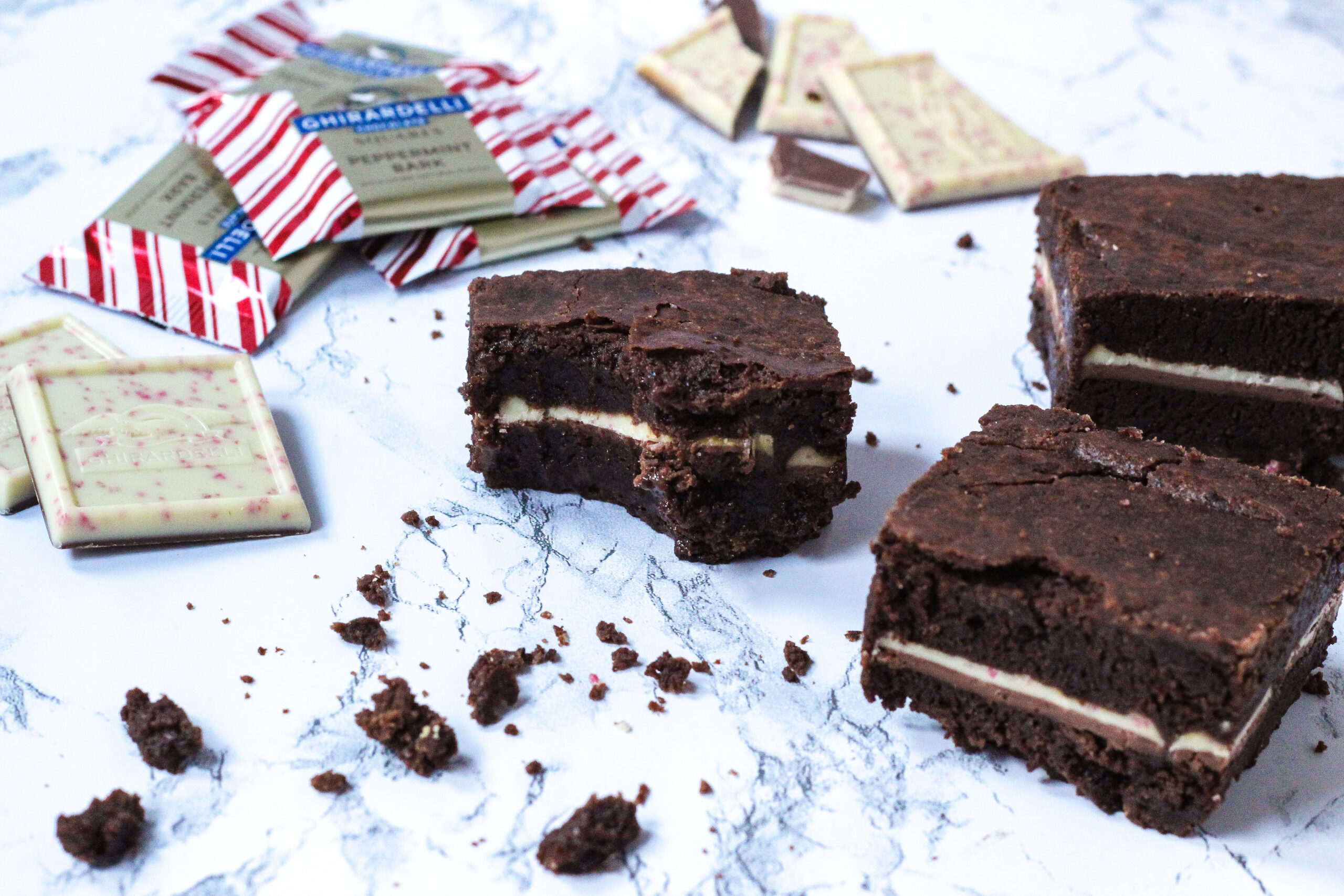 angled view, with a brownie with a bite taken out of it in the center of the frame. to the left of the brownie is whole, unwrapped peppermint bark squares, some wrapped peppermint bark squares in the top left corner. Top center, behind the brownie, is broken pieces of peppermint bark squares. Along the right side of the frame is two more brownies. In front of the bitten brownie at the bottom of the frame is a bunch of brownie crumbs.