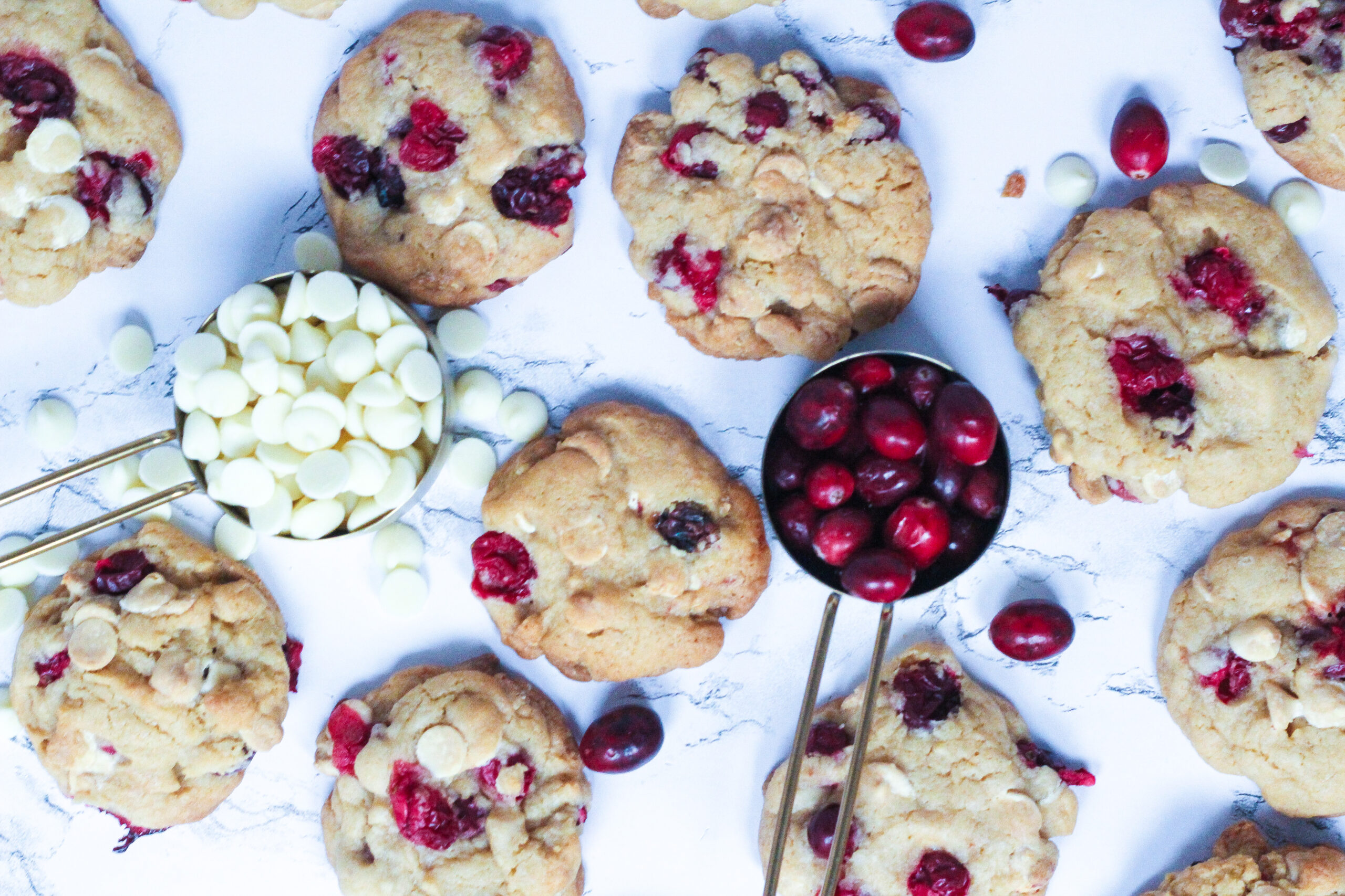 top down view of cookies scattered around a white marble surface, with a gold measuring cup in center left of the frame with white chocolate chips and another center right with cranberries.