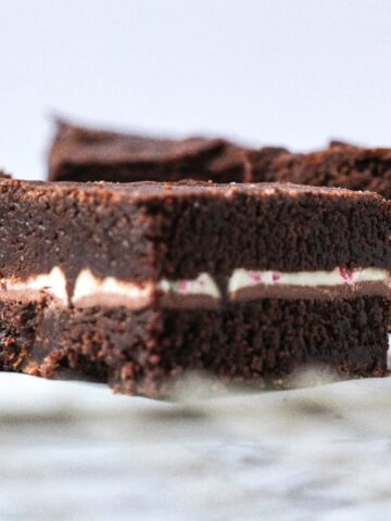 Close up side view of a peppermint mocha brownie, with other peppermint mocha brownies blurred in the background. The brownie in focus is at an angle so the corner is facing the camera. The peppermint mocha brownies are dark brown brownies with a thin white layer in the center from the peppermint bark square