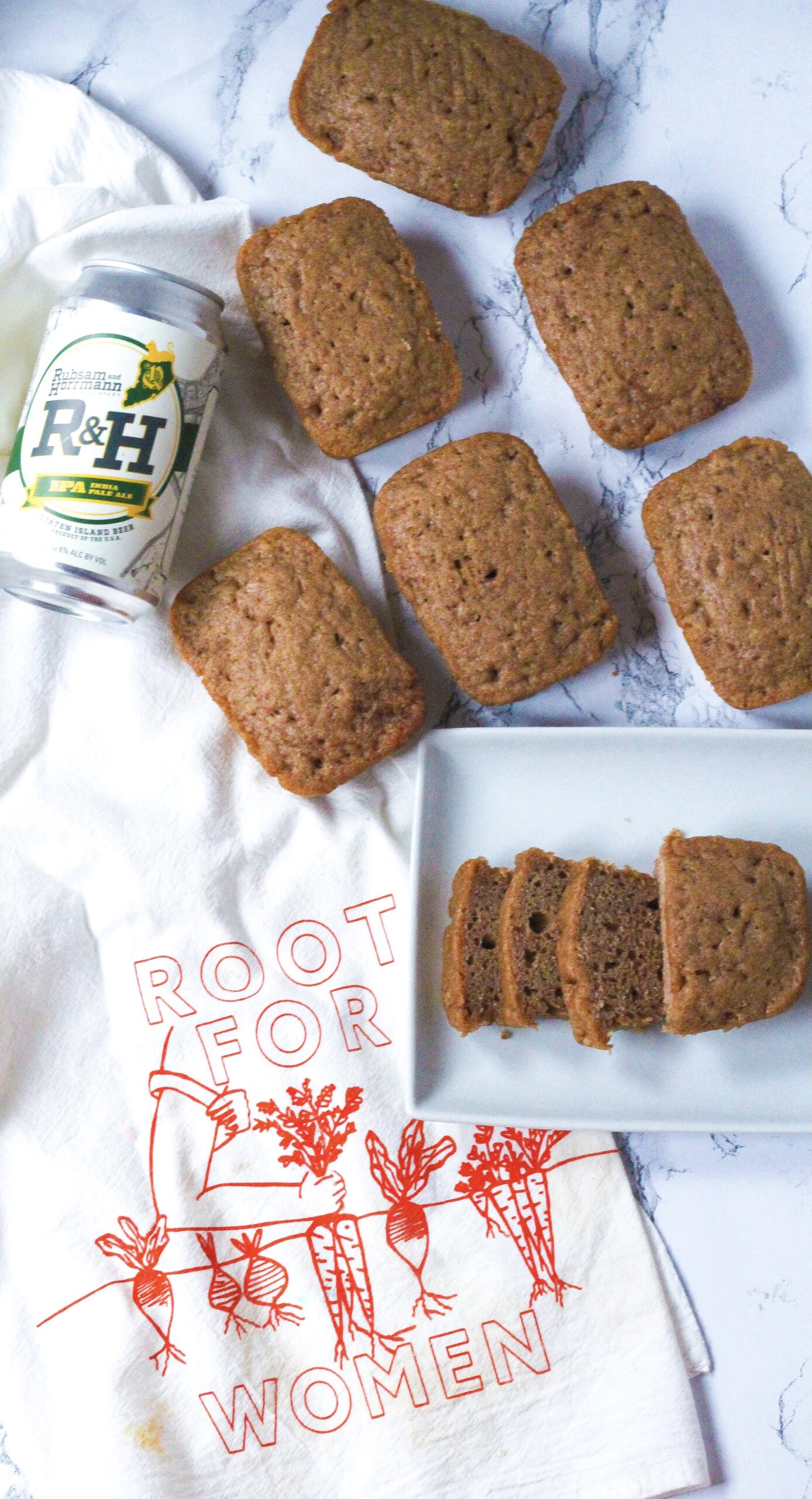 Top down view of, on the left side of the photo is a white dish towel that says Root for Women in red with root vegetables on it. In the upper left of the image on top of the towel above the words are some mini loaves of spiced beer bread and a can of R&H IPA Beer. On the right side of the photo is a white rectangular plate that slightly overlaps the towel. On the plate in the back is a mini loaf, and in the front is a loaf, the front half of which is sliced into three slices.